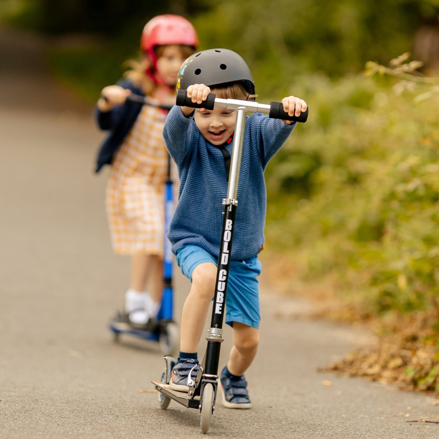 Two children riding scooters on a path with greenery in the background