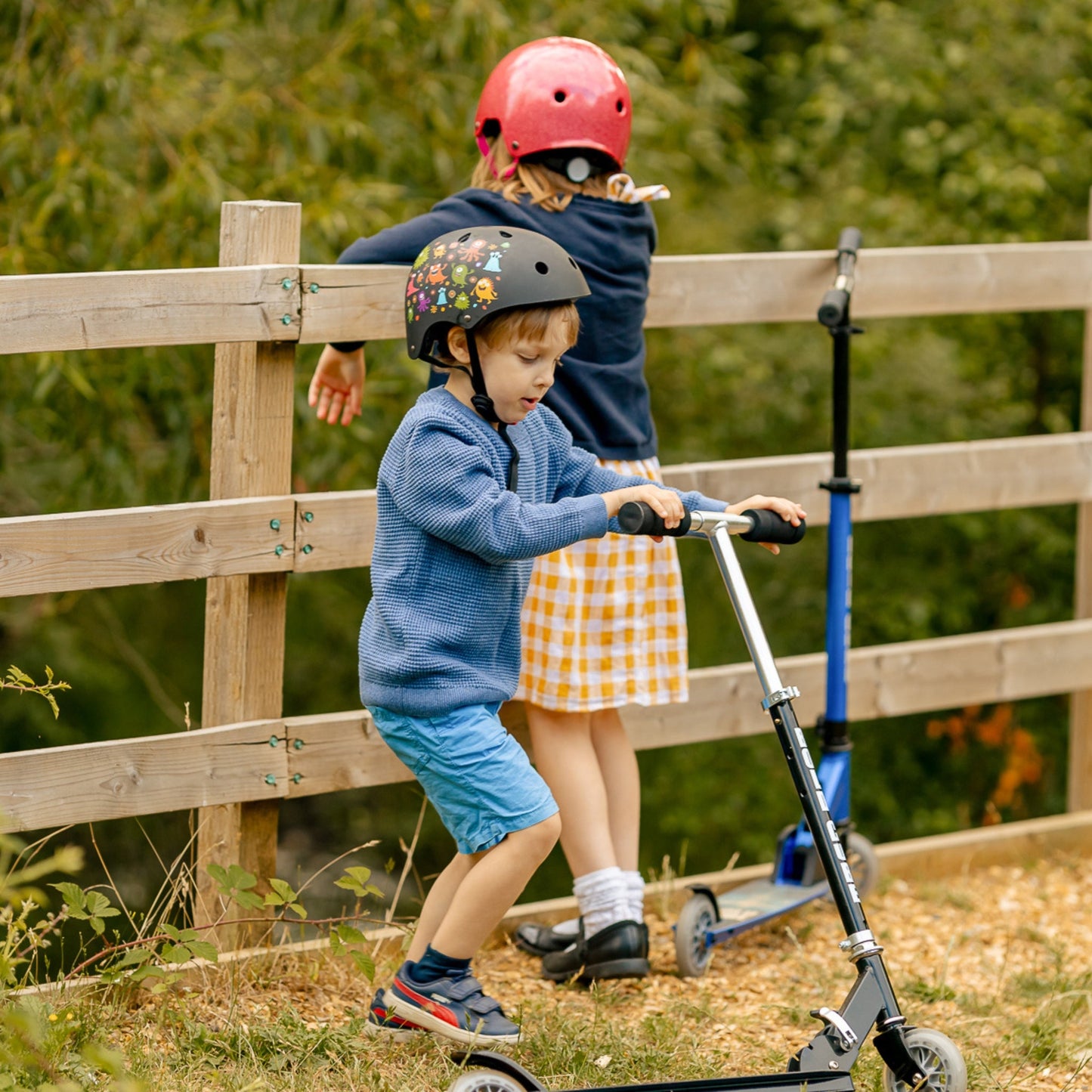 Two children on scooters with helmets near a wooden fence.