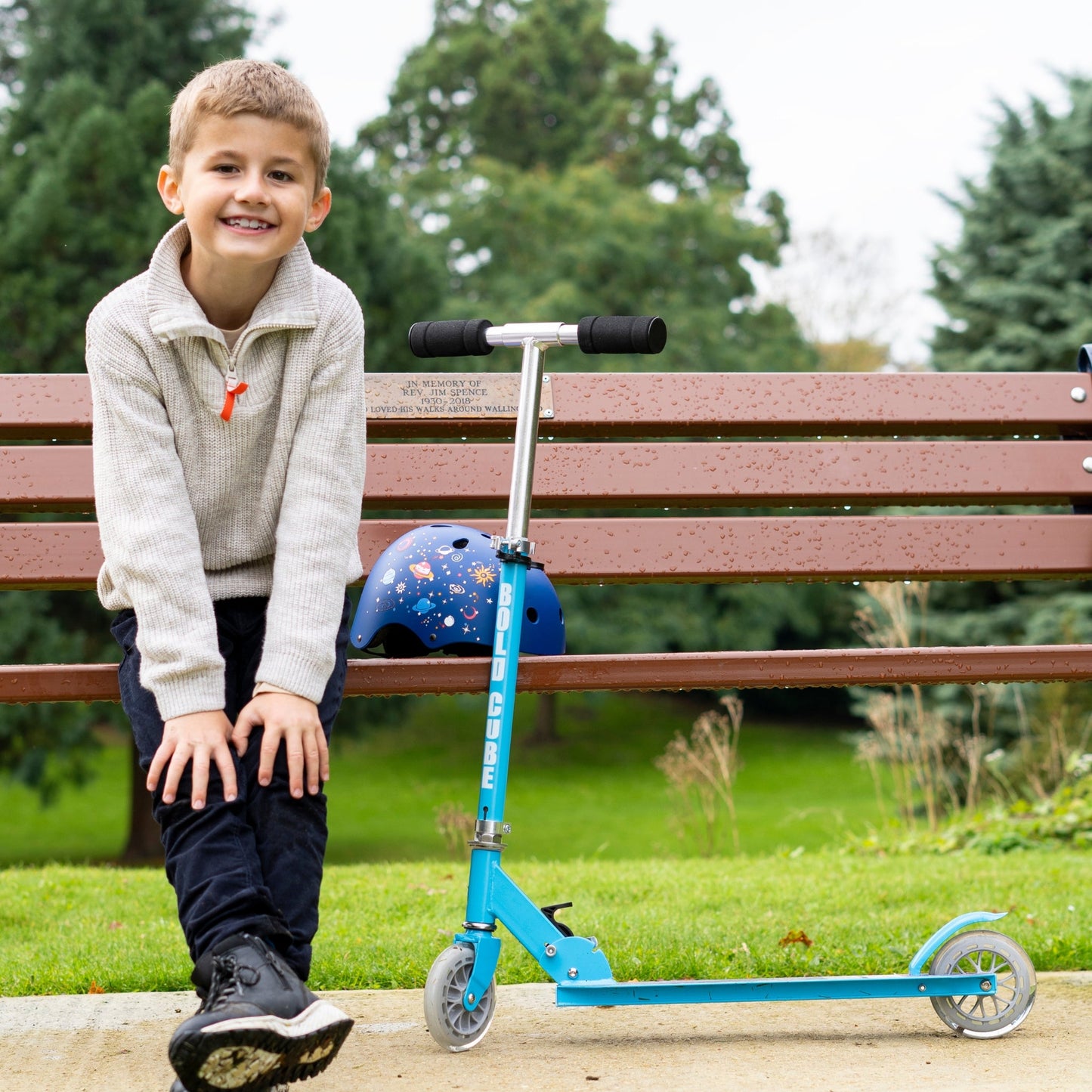 Child sitting on a bench with a blue scooter and helmet in a park setting