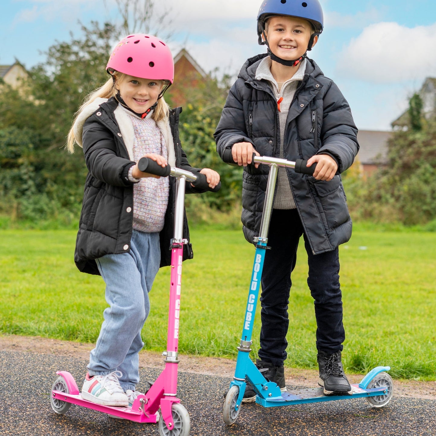 Two children on scooters wearing helmets in a park setting