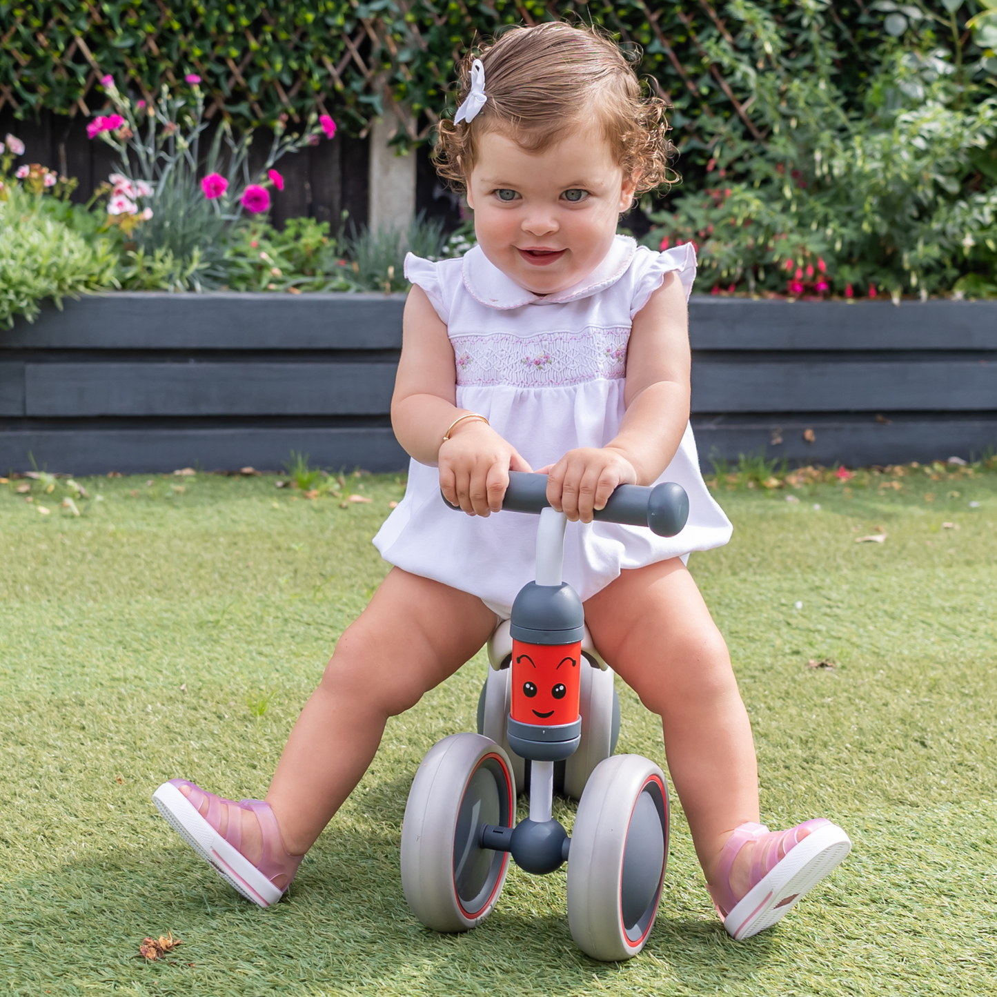 Child riding a toy bike outdoors on grass with flowers and plants in the background