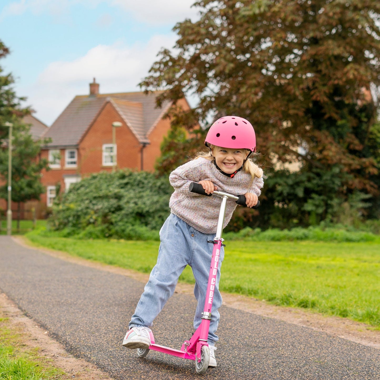 Child riding a pink scooter on a residential street with houses and greenery in the background.