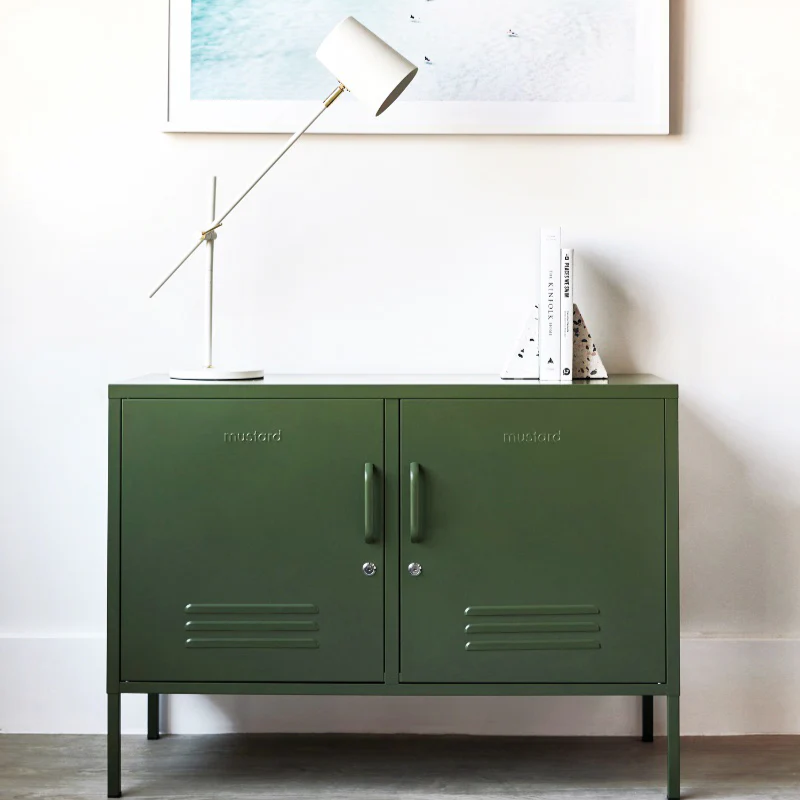 Green metal cabinet with branding, lamp, and abstract painting on a white wall.