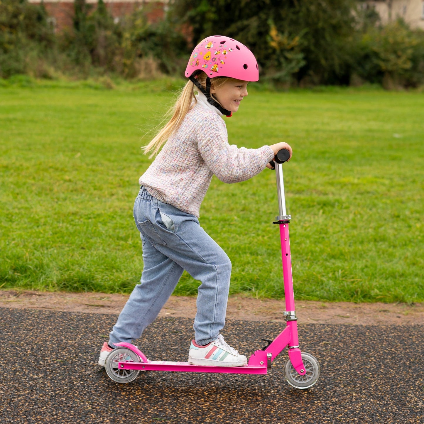 Child riding a pink scooter on a paved path with grass and trees in the background