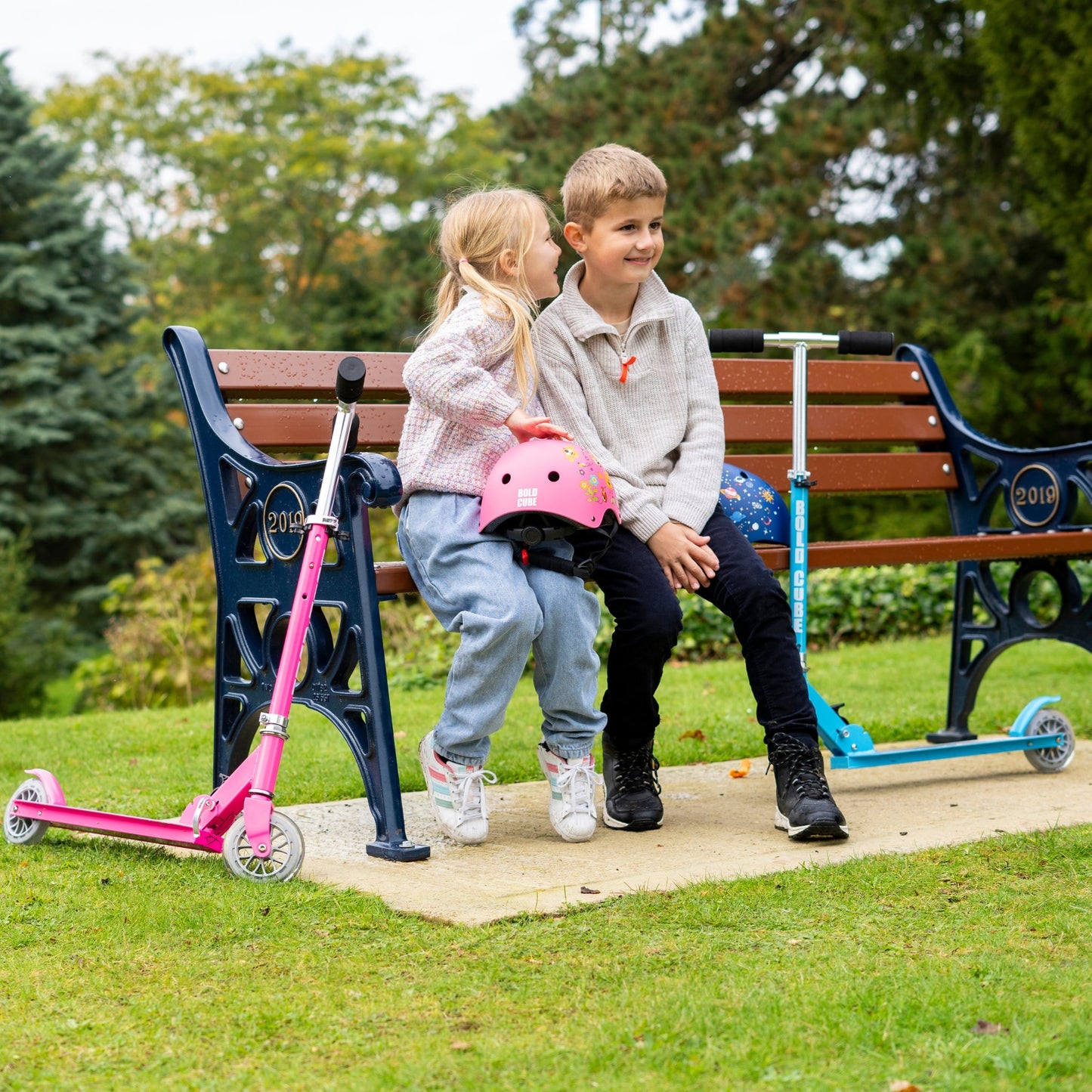 Two children with scooters sitting on a park bench