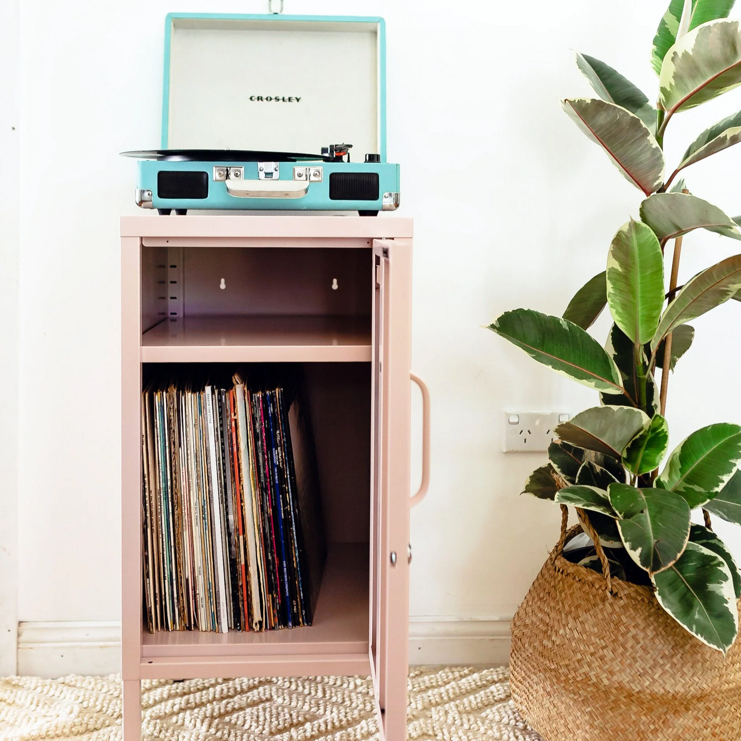 Record player on a pink cabinet with vinyl records, a plant, and colorful abstract art in the background.