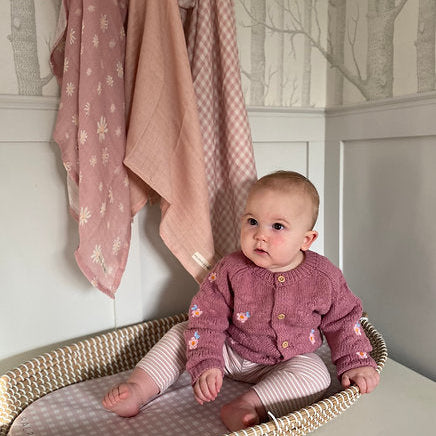 Baby in a pink cardigan sitting in a crib with pink blankets in the background