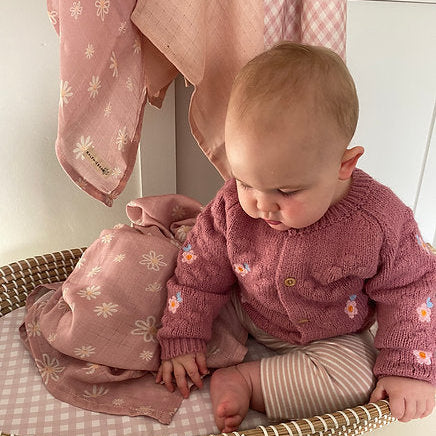 Baby in pink outfit sitting on a woven basket with pink clothing in the background