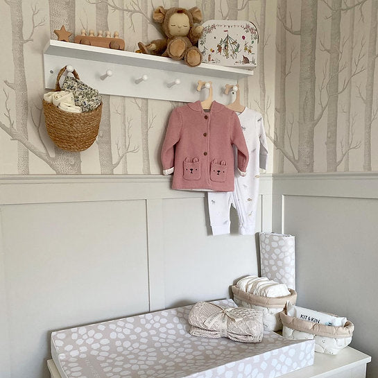 Nursery setup with changing table, baby clothes, and toys against a tree-patterned wall.