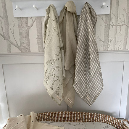Three baby blankets hanging on a hook against a wall with tree wallpaper.