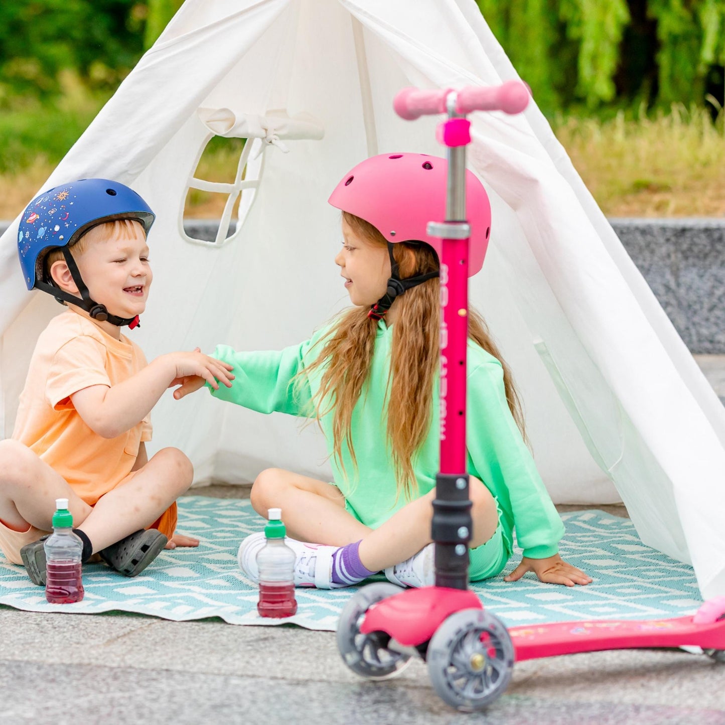 Two children with helmets sitting on a blanket next to a pink scooter in front of a white tent.