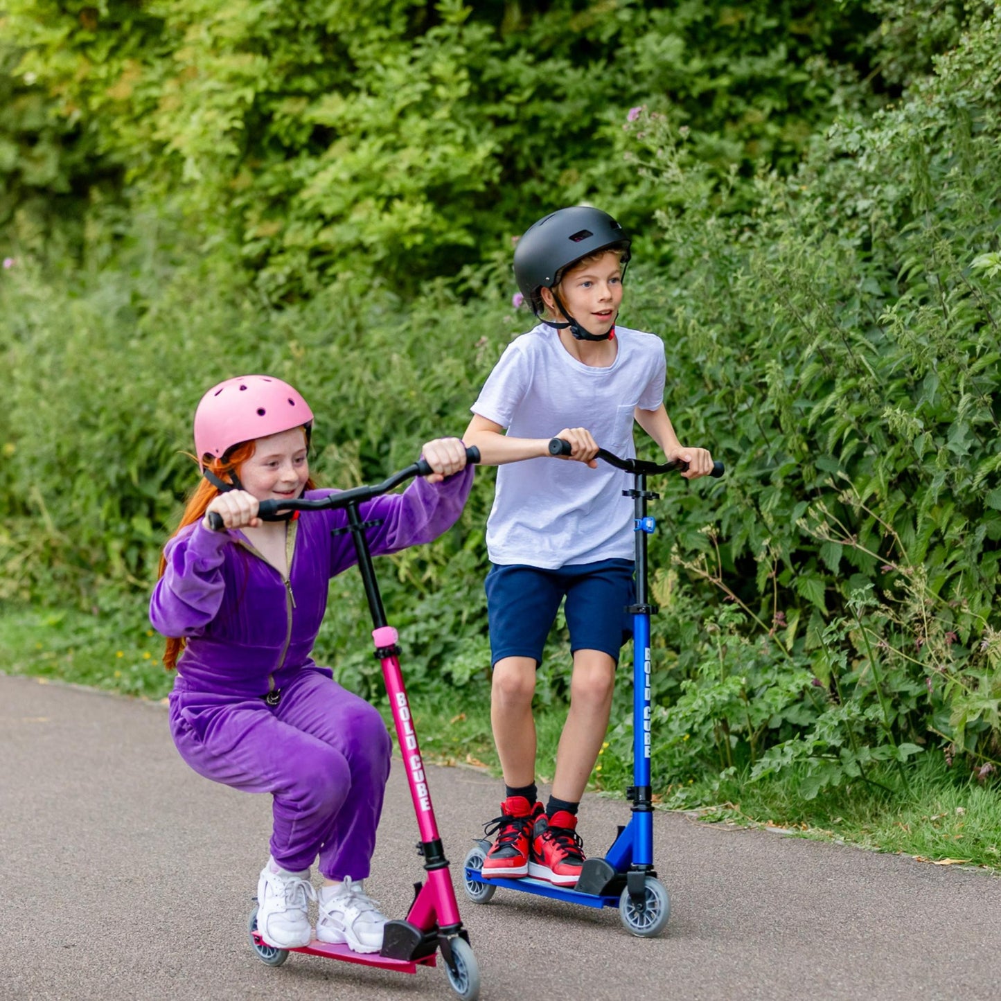 Two children riding scooters on a path with greenery in the background