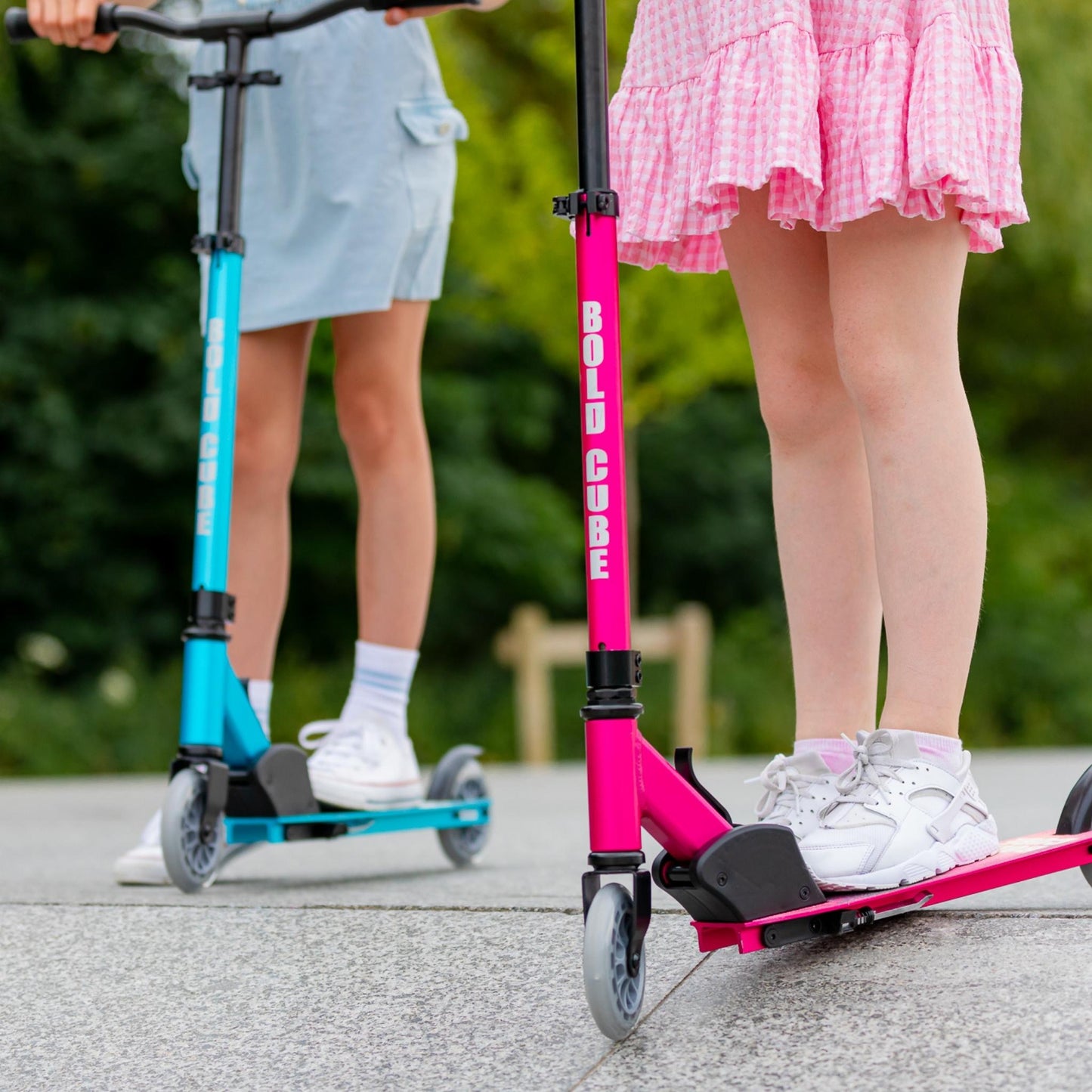 Two children on scooters, one pink and one blue, with a blurred green background.