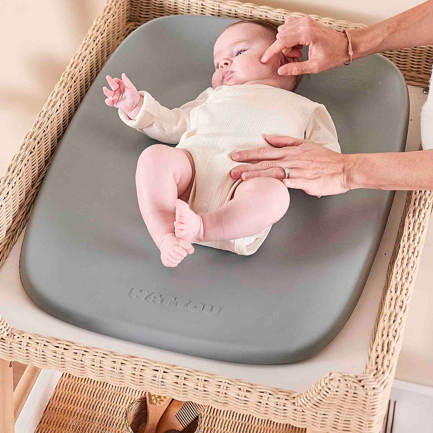 Baby sitting on a gray changing pad with a person's hands near, on a wicker chair.