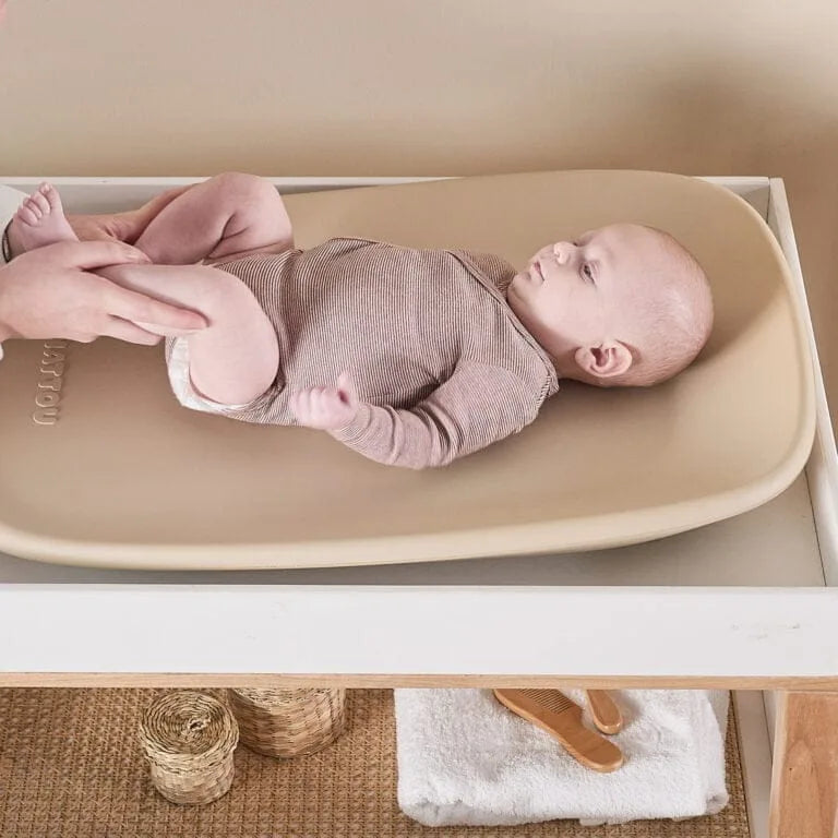 Baby lying on a changing table with a person's hand, set against a neutral background.
