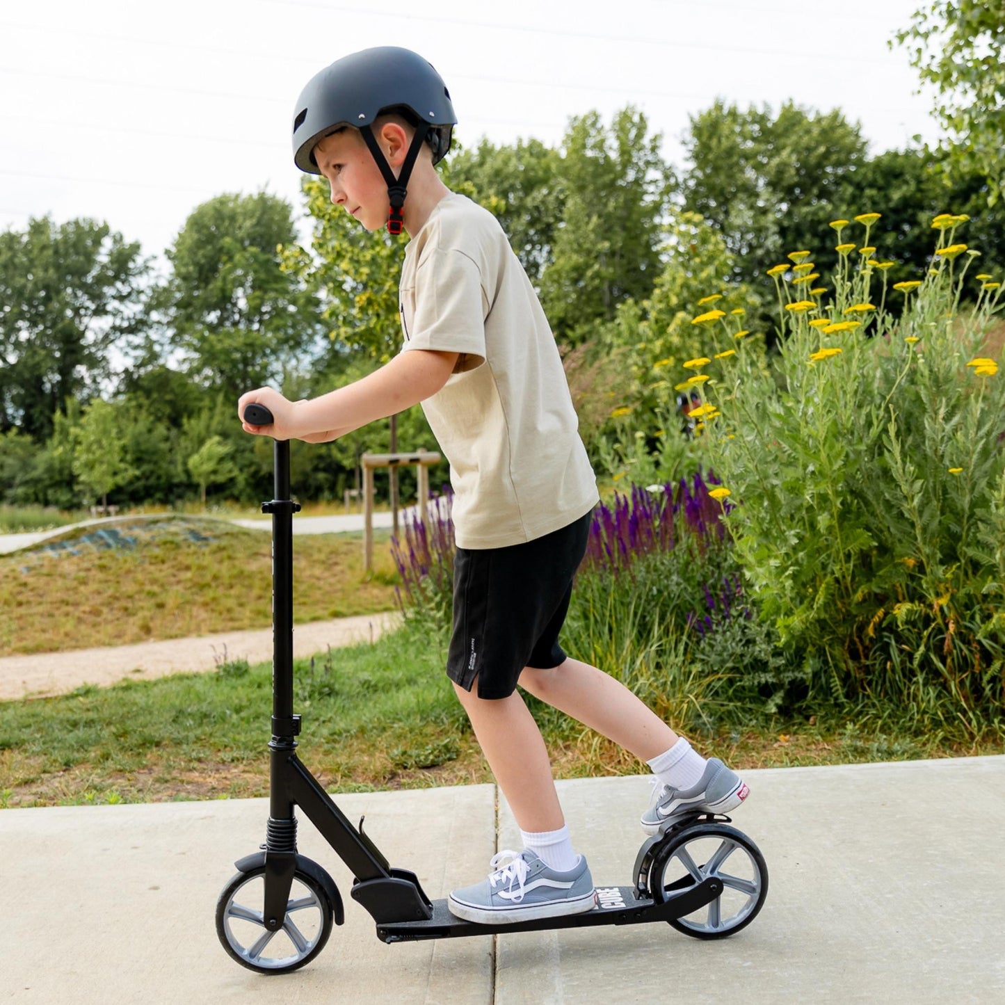 Child riding a scooter in a park with trees and flowers in the background