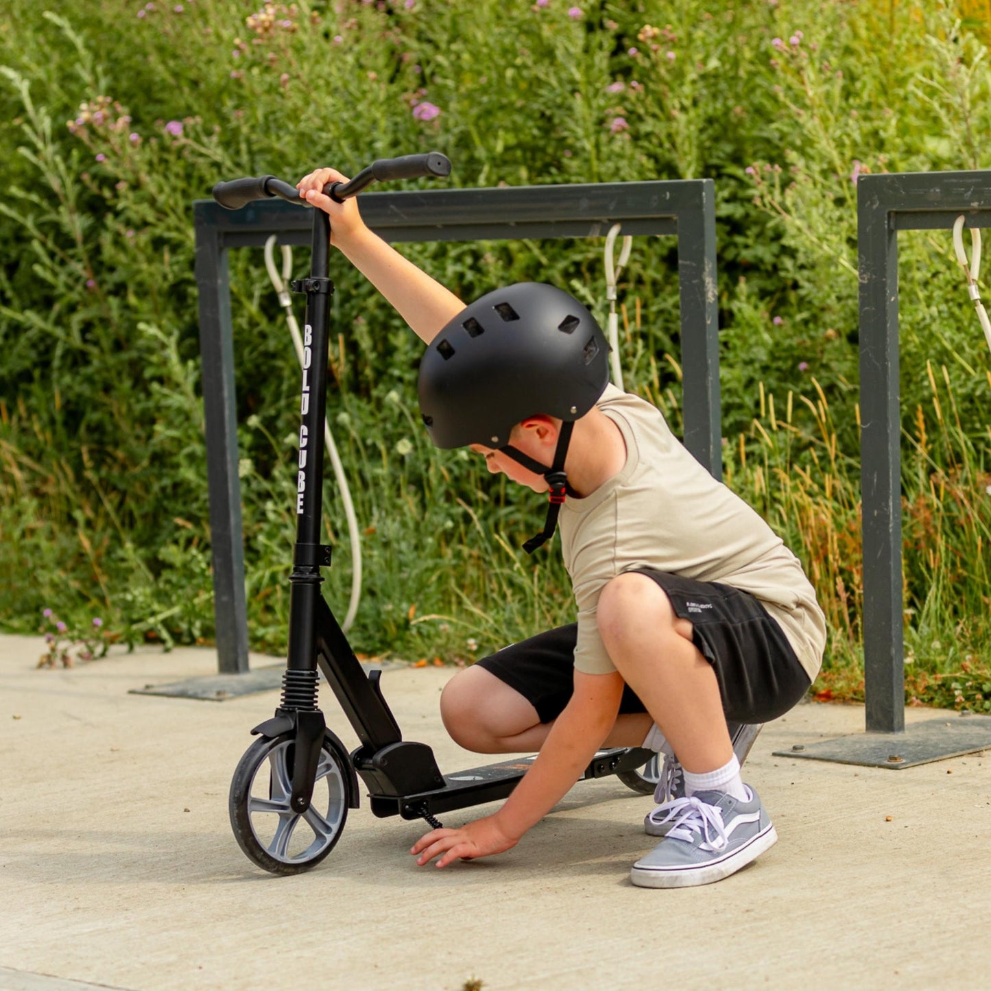 Child with a scooter outdoors on a wooden path with greenery in the background