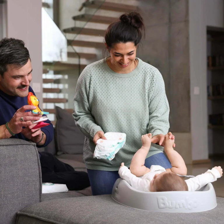 Woman changing a baby's diaper in a living room with a man holding a toy.
