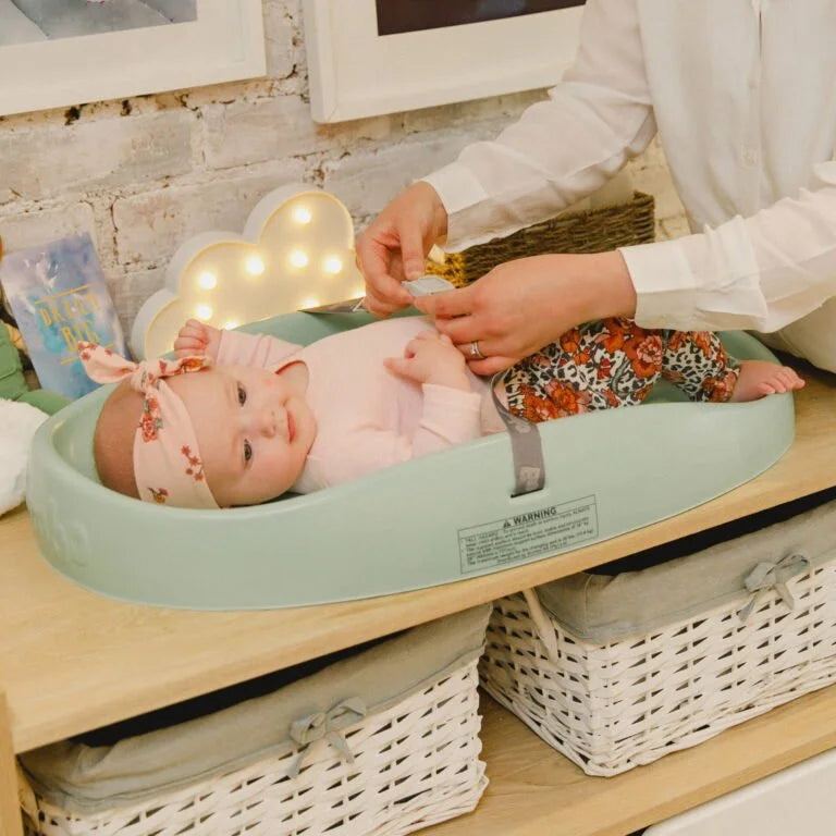 Baby lying in a green bassinet with a person adjusting something above, in a home setting.