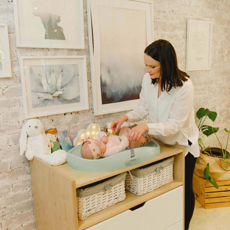 Woman changing a baby on a changing table in a home setting with framed artwork and plants.