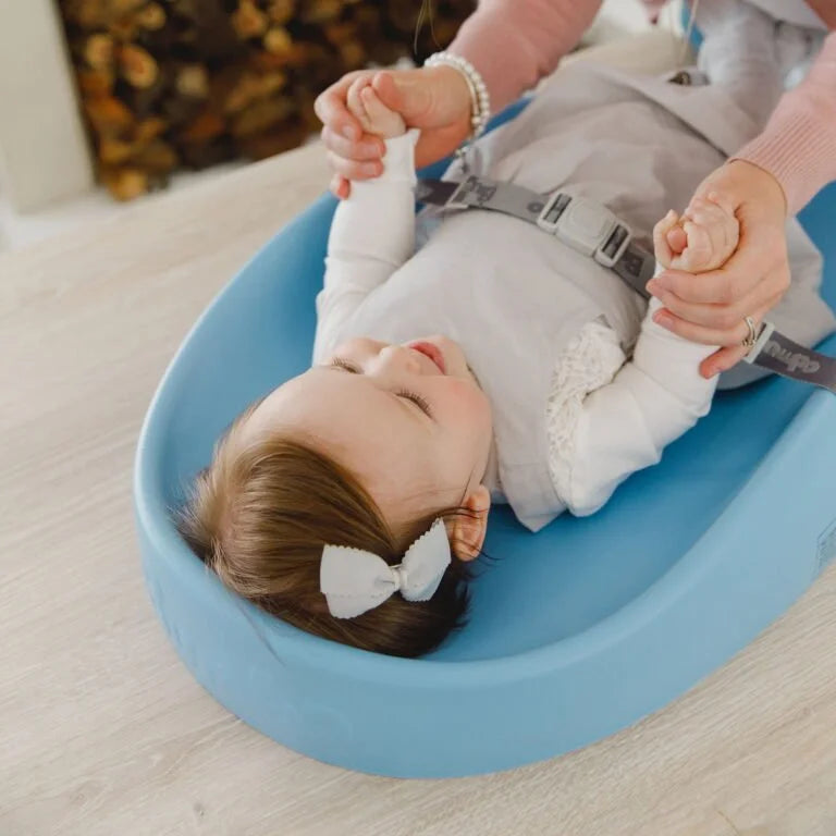 Baby lying on a blue bath seat with adult hands holding its feet, on a light wooden floor.