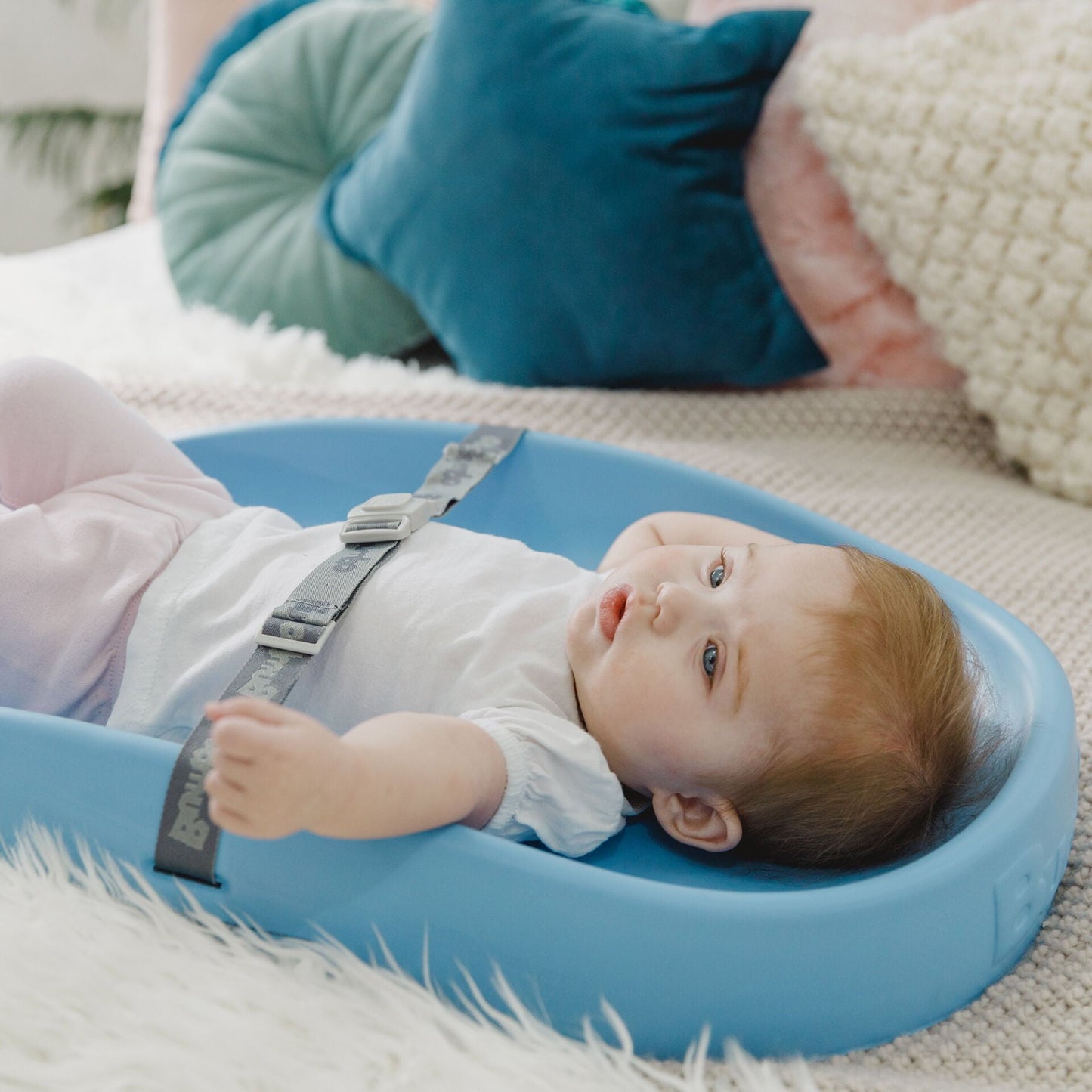 Baby lying on a blue mat with straps, surrounded by cushions on a couch.