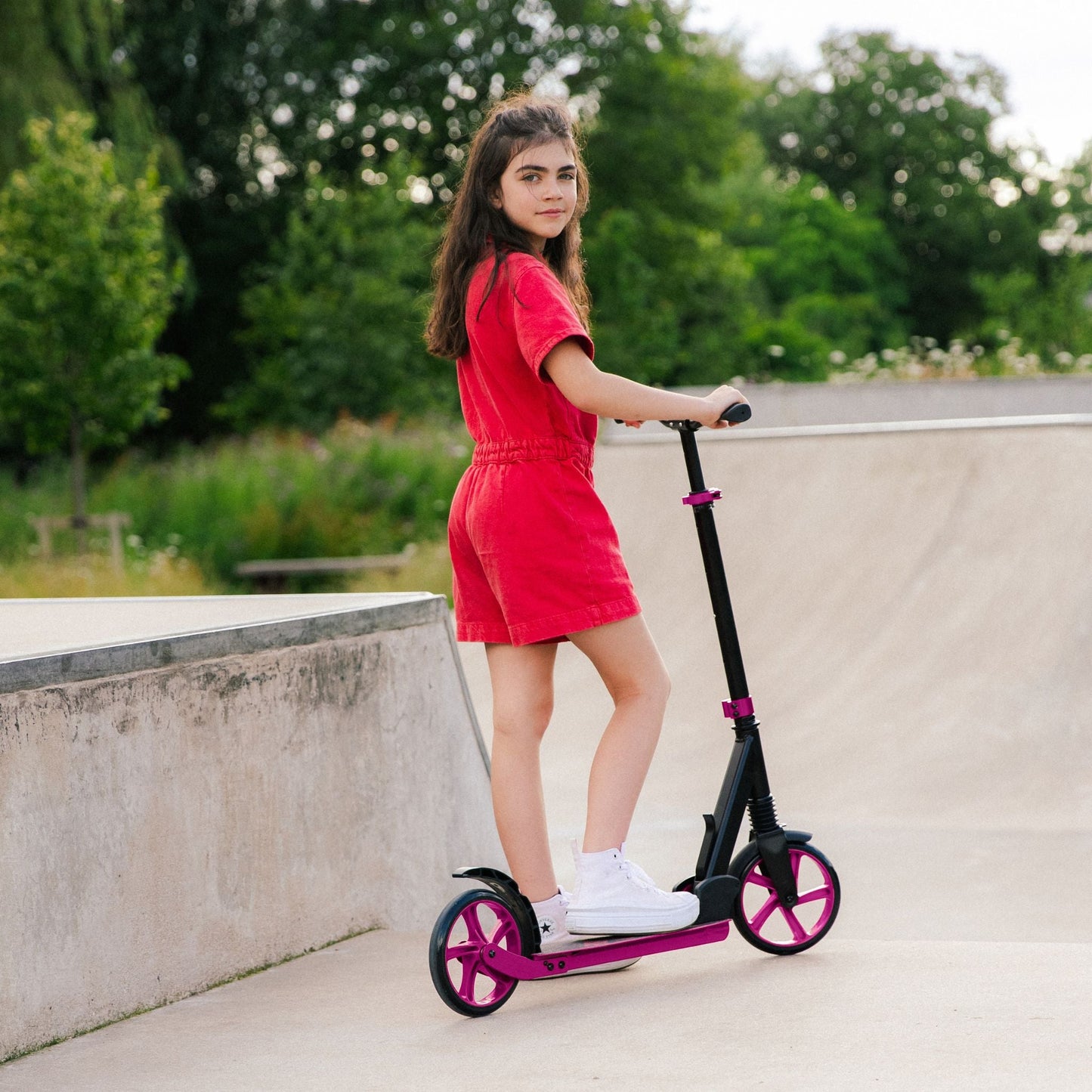 Girl in a red dress standing on a scooter at a skate park.