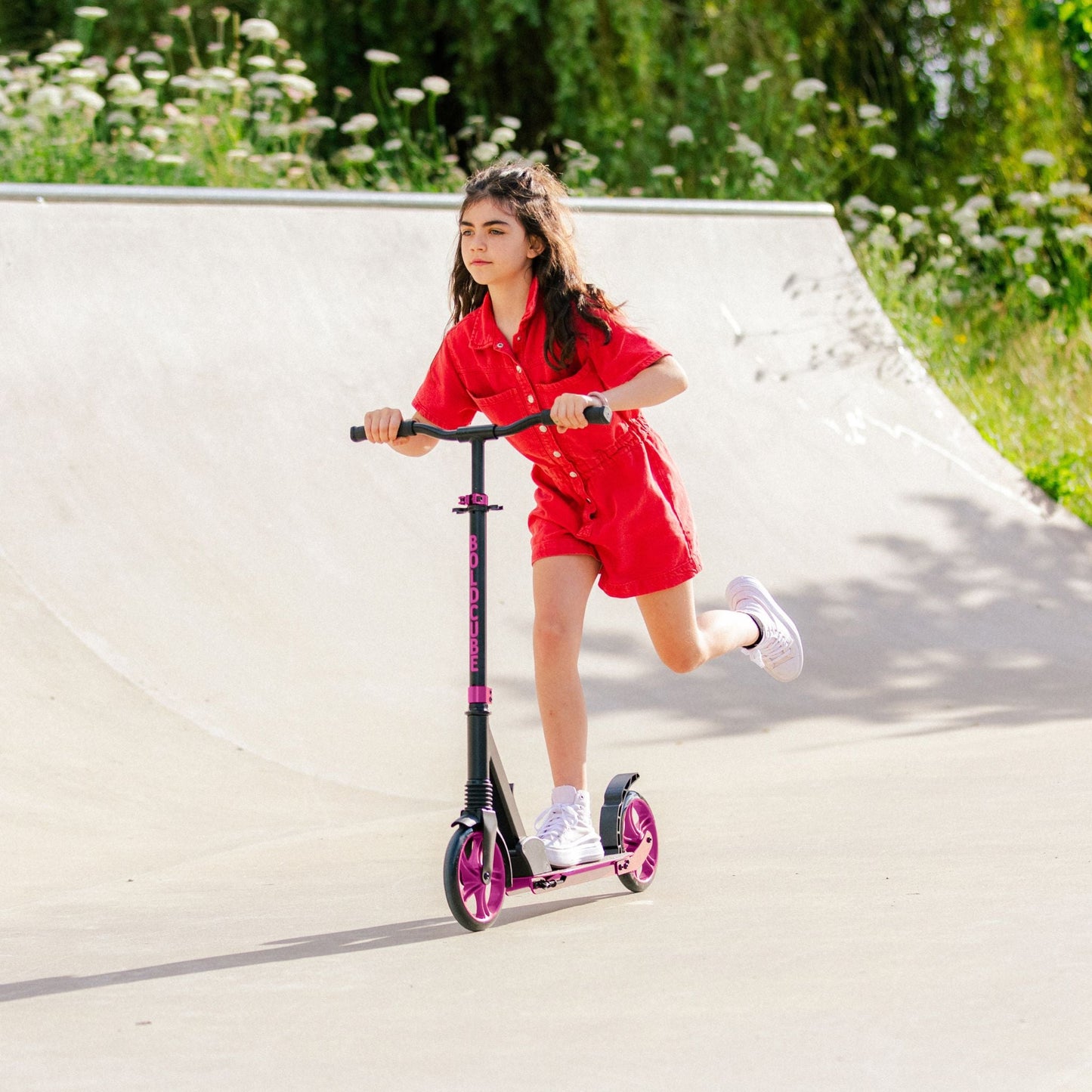 Girl in red outfit riding a scooter on a paved path with greenery in the background