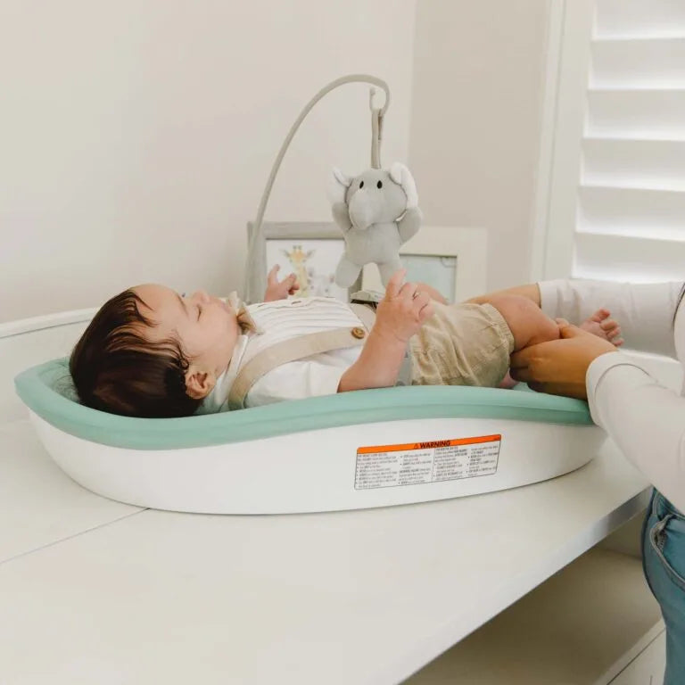 Baby lying on a green and white baby seat with a plush toy in the background