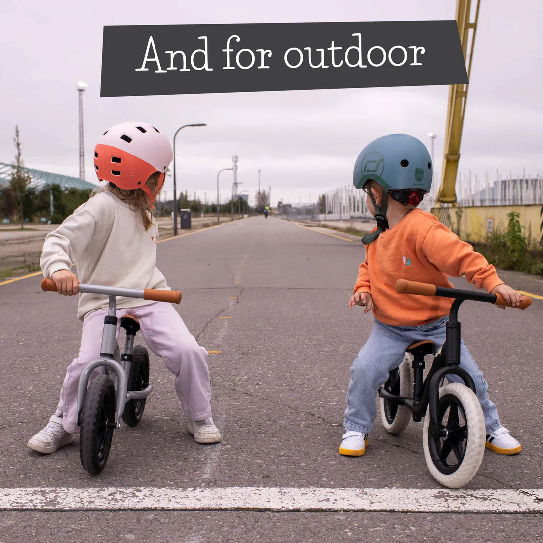 Two children riding balance bikes on an outdoor road with helmets on.