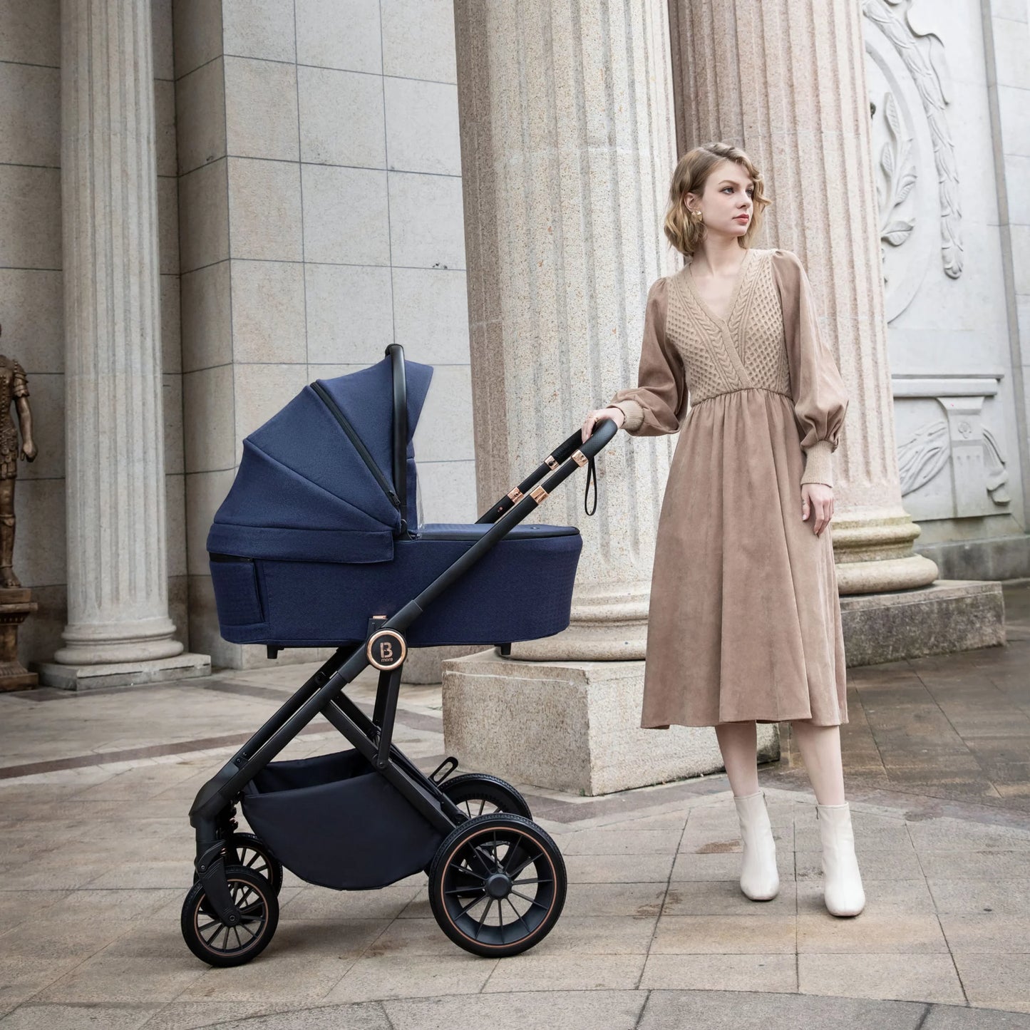Woman in a beige dress standing next to a navy blue stroller in an architectural setting.