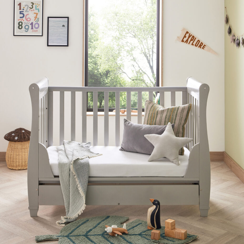 Gray crib with bedding and toys in a room with large windows and decor.