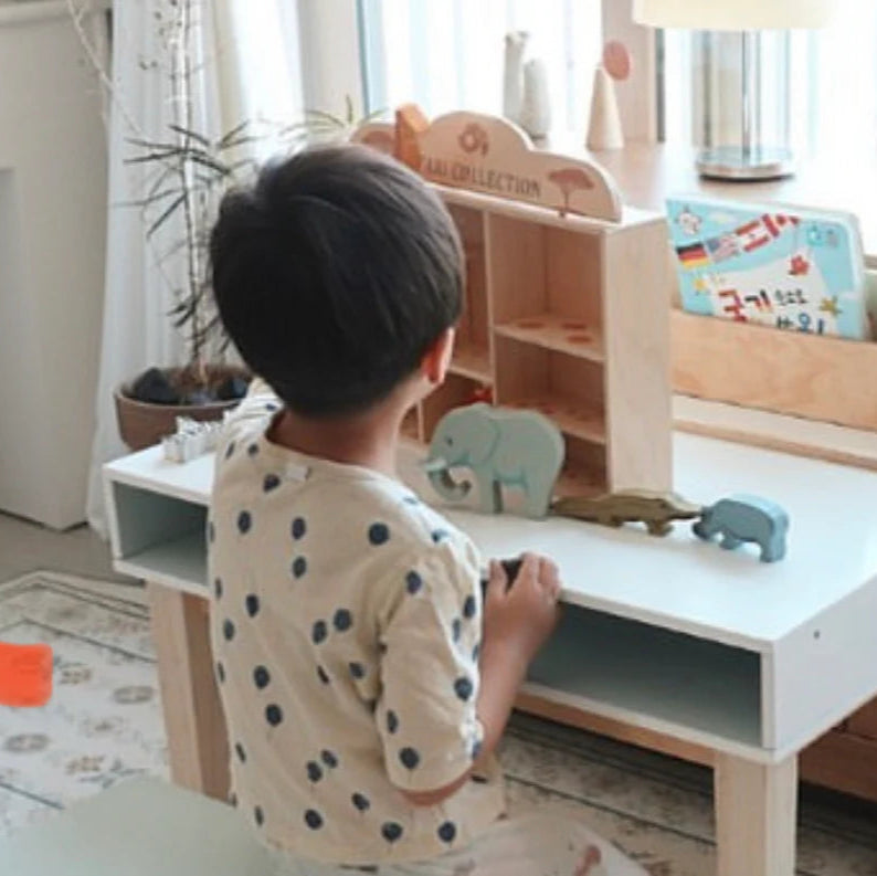 Child playing with toys on a small wooden desk in a bright room.