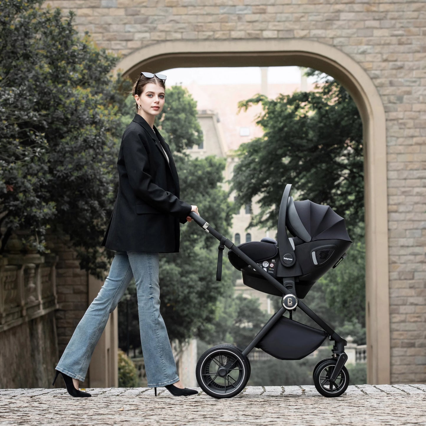 Woman pushing a black stroller in an outdoor setting with a stone archway and greenery.