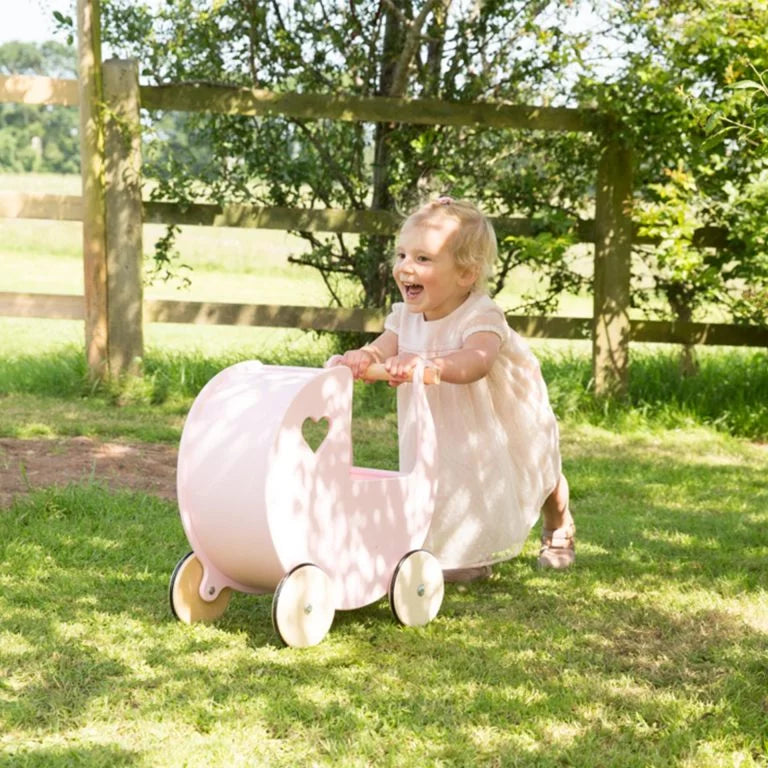 Child in a white dress playing with a pink toy cart in a grassy outdoor setting.
