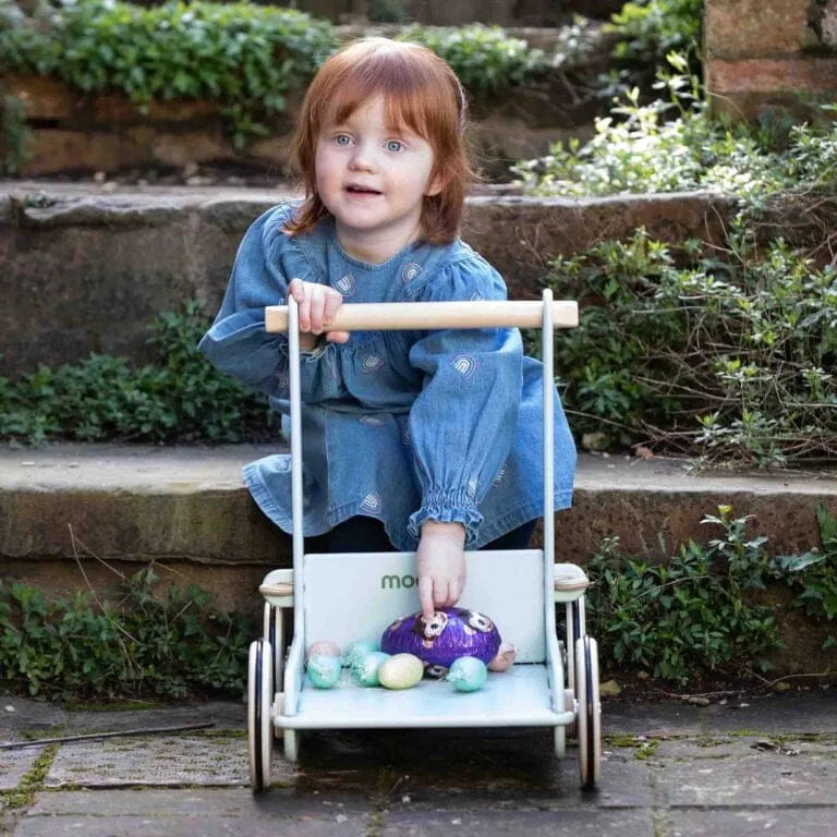 Child sitting in a toy stroller with a purple teddy bear and colorful eggs outdoors.