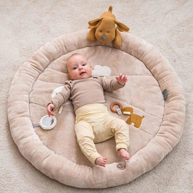 Baby lying on a round beige mat with toys on a light gray background
