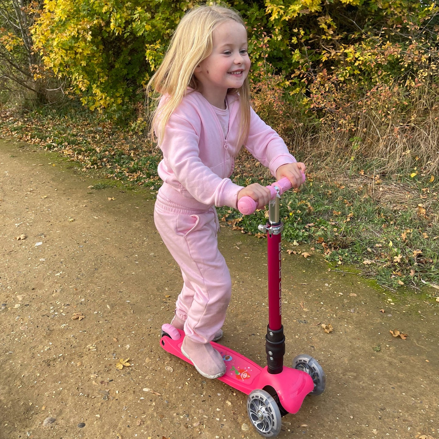 Child in pink outfit riding a pink scooter outdoors with trees in the background