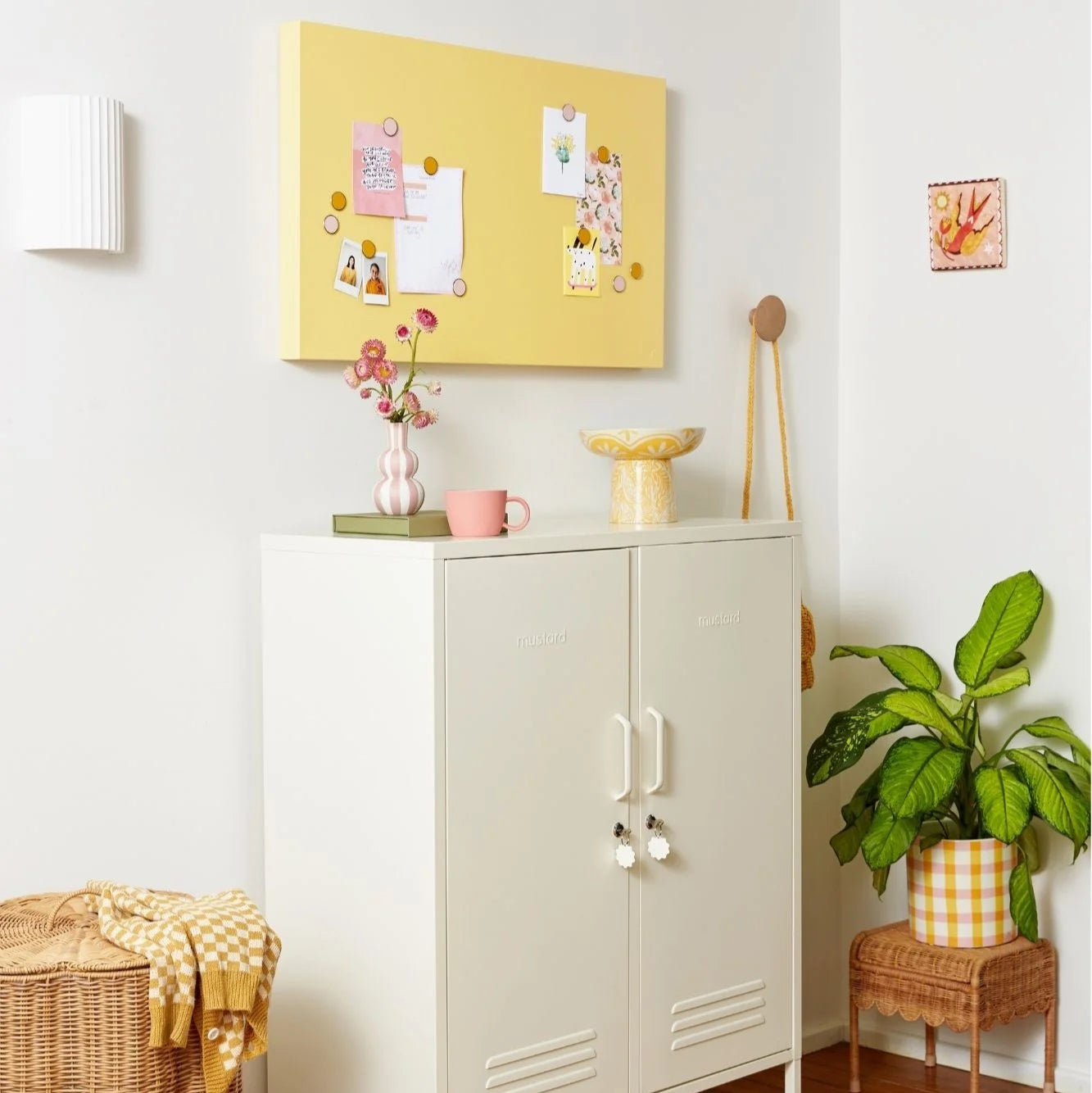 White cabinet with a yellow bulletin board above it in a room with plants and a basket.