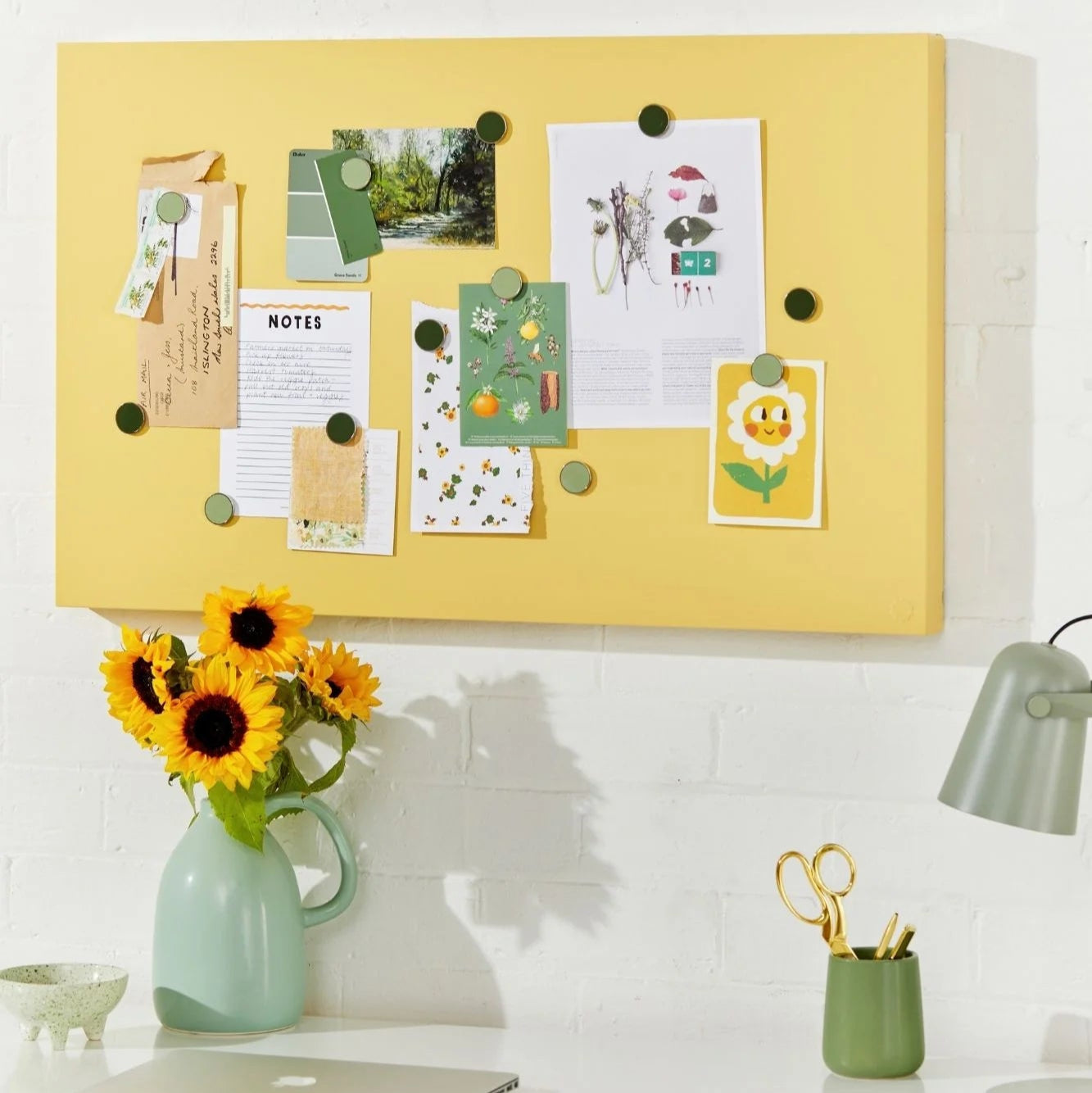 Yellow bulletin board with pinned notes above a desk with a vase of sunflowers and office supplies.