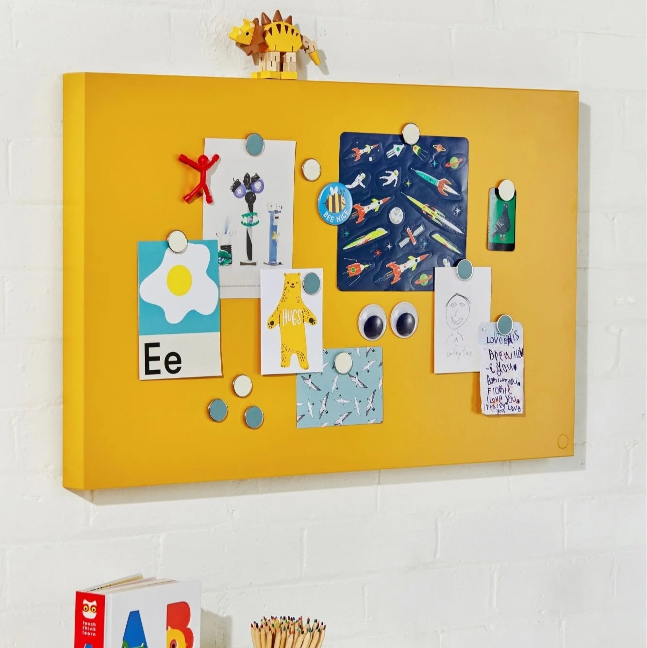 Yellow magnetic board with children's drawings and letters on a white wall.