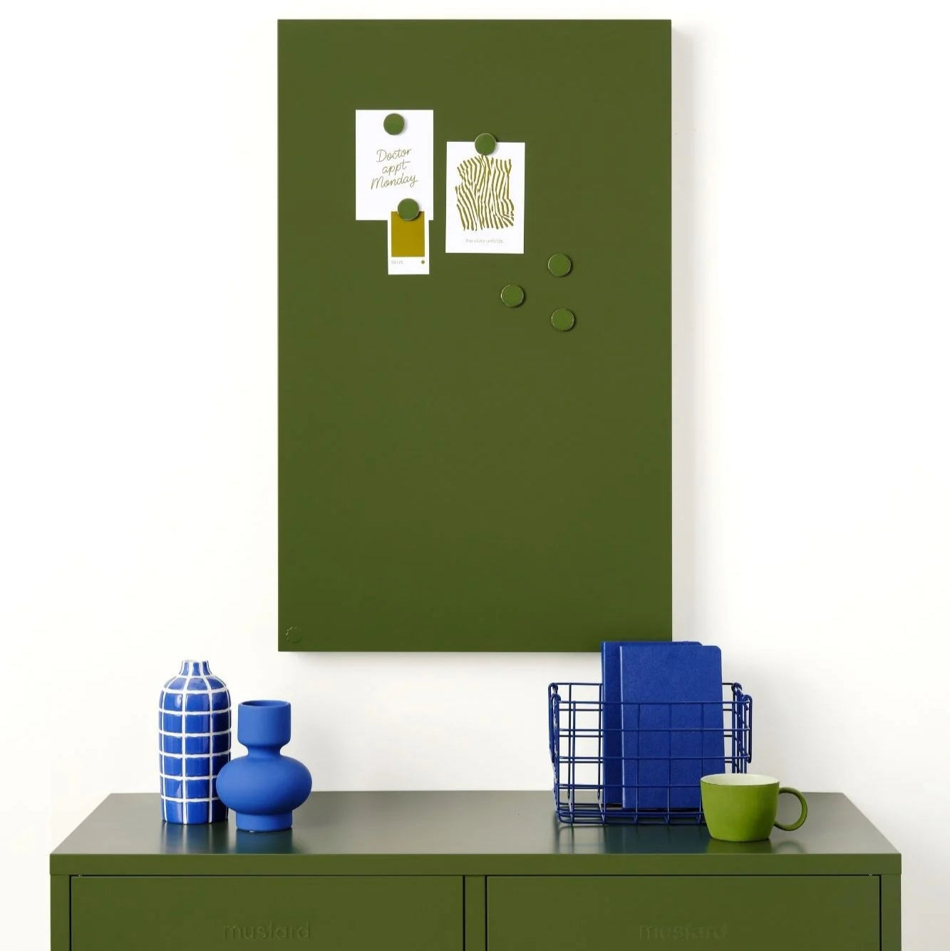 Green magnetic board on a white wall above a green cabinet with decorative items.