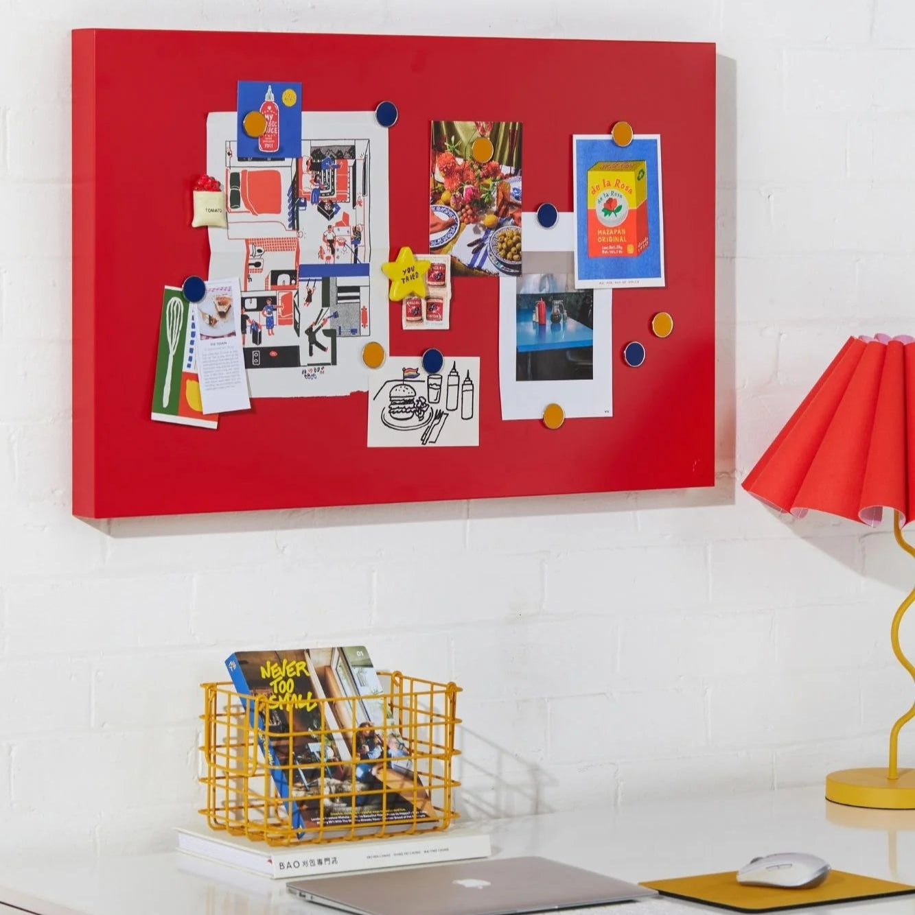 Red bulletin board with photos and notes on a white wall above a desk with office supplies.
