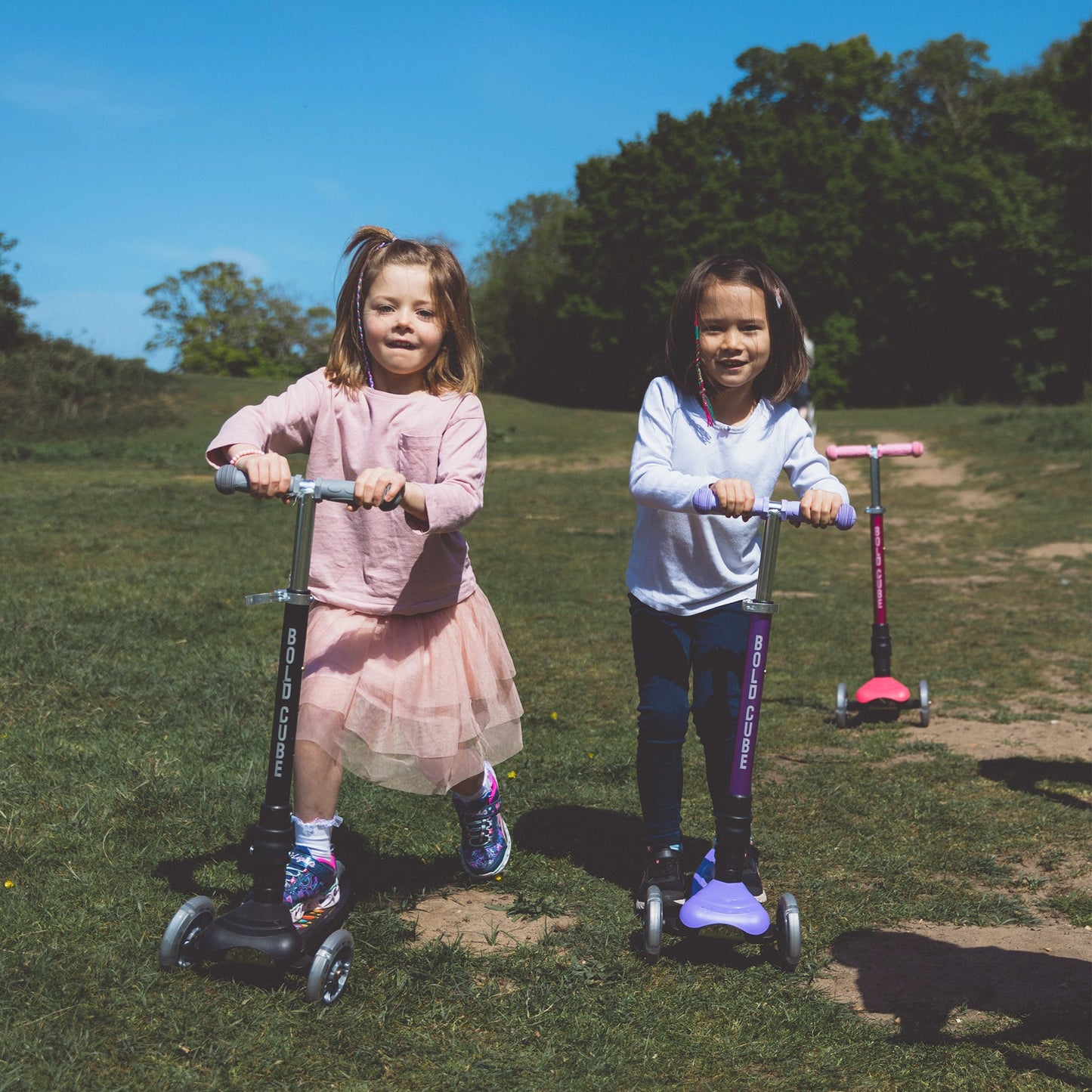 Two young girls on scooters in a park with trees and grass.