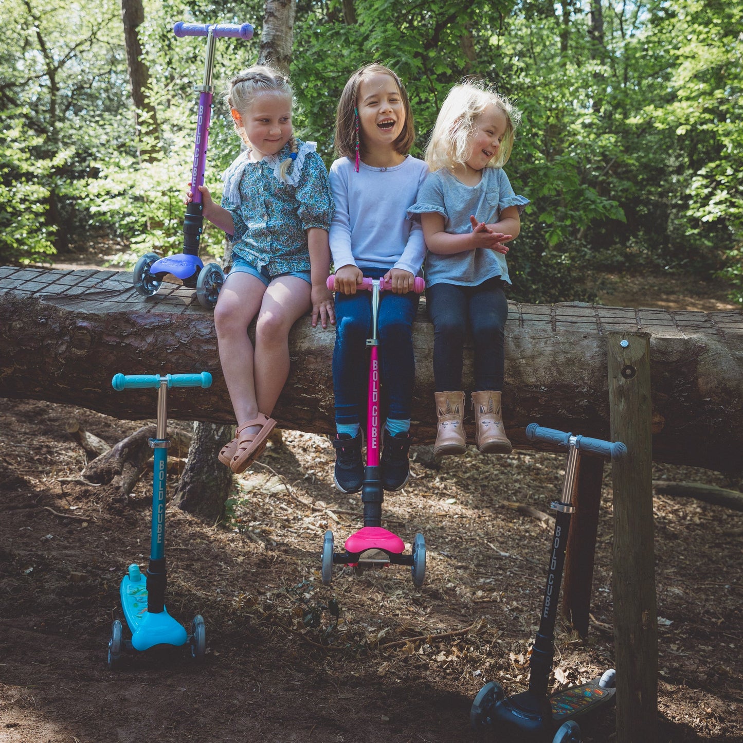 Three children with scooters on a log in a forest setting