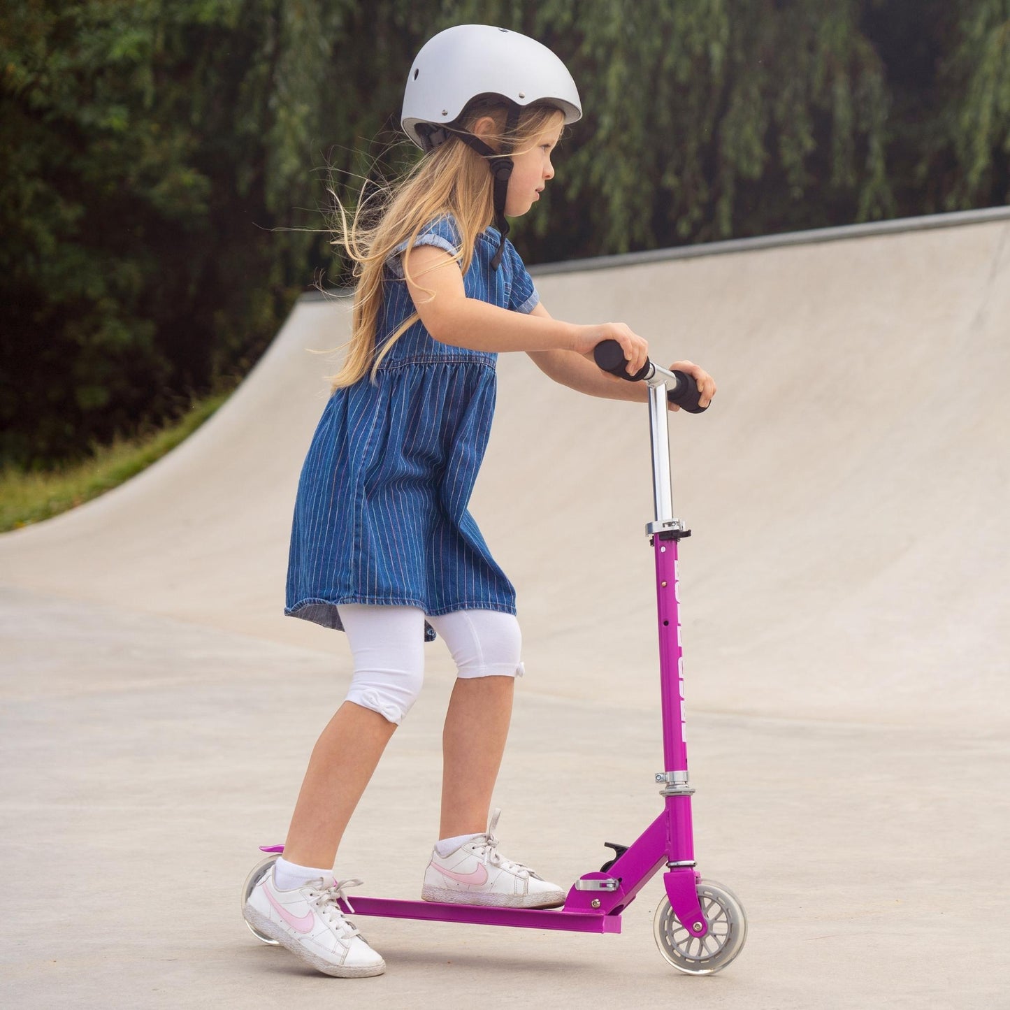 Child riding a pink scooter on a concrete surface with greenery in the background