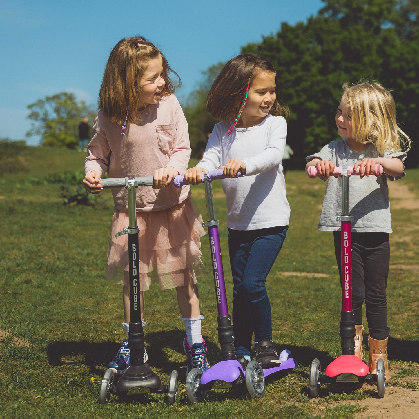 Three children on scooters in a grassy outdoor setting
