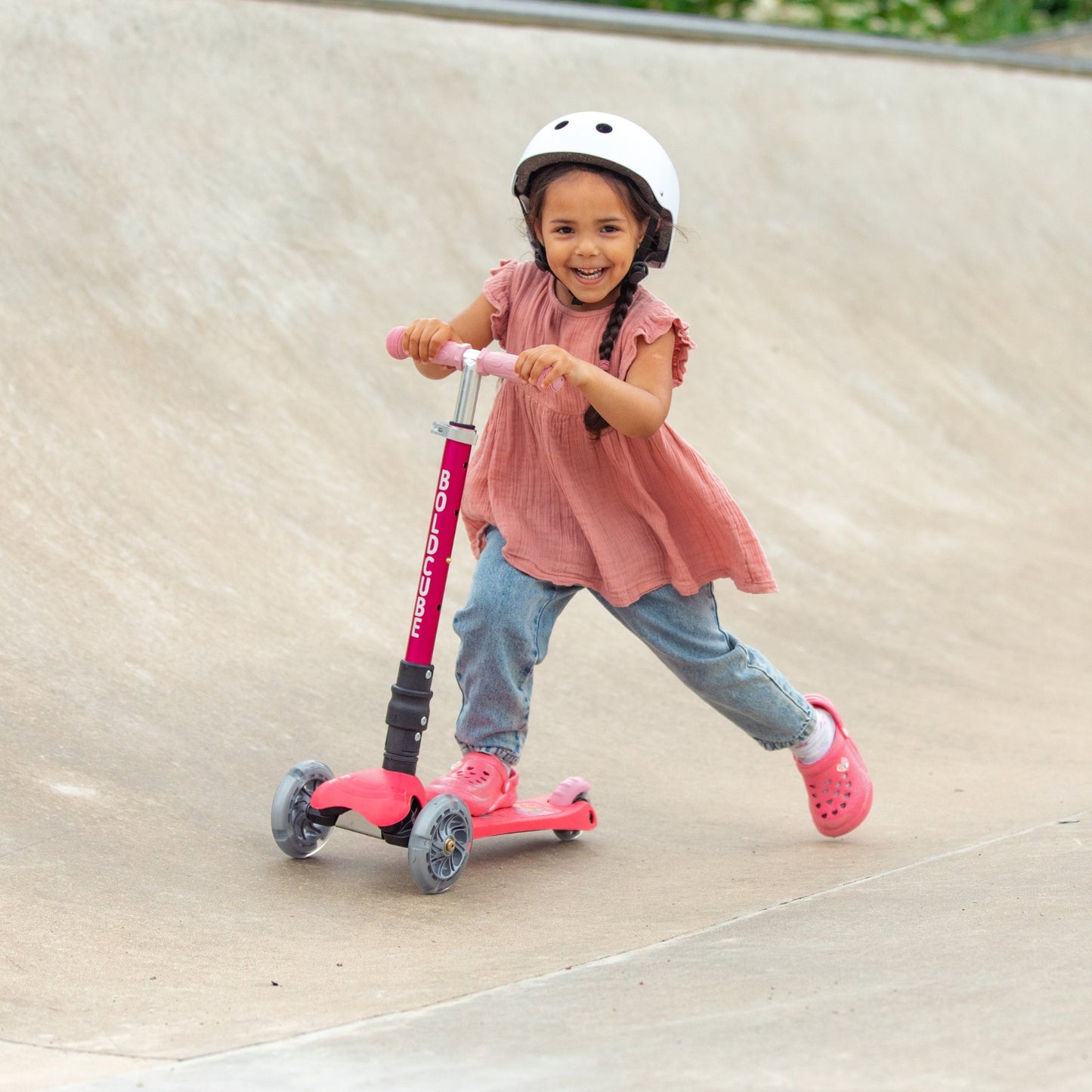Child riding a pink scooter with a helmet on a concrete surface