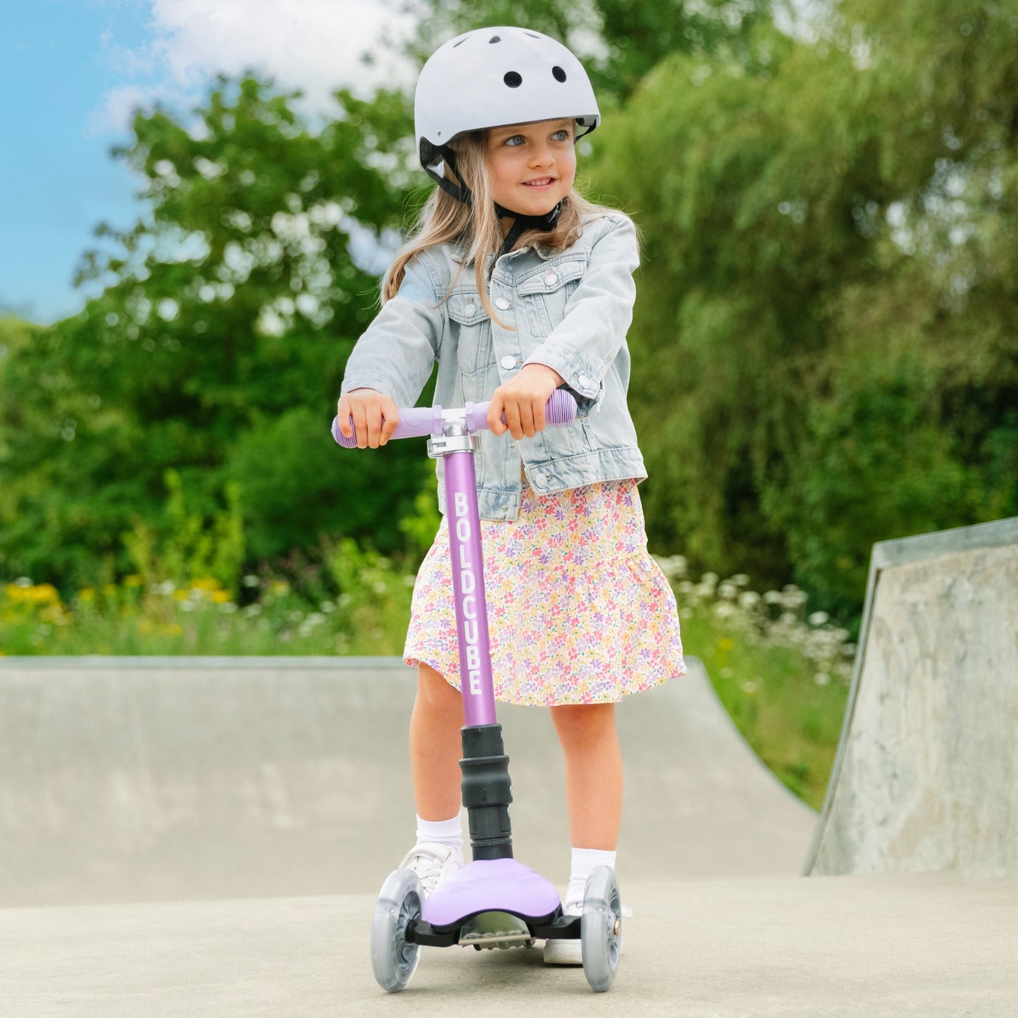 Child riding a purple scooter with a helmet on a paved path with greenery in the background