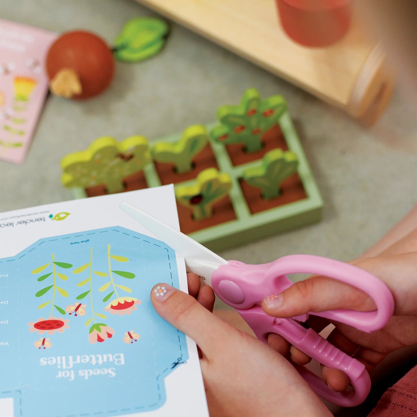 Child's hand holding pink scissors near a book with a garden illustration on a table with toys.